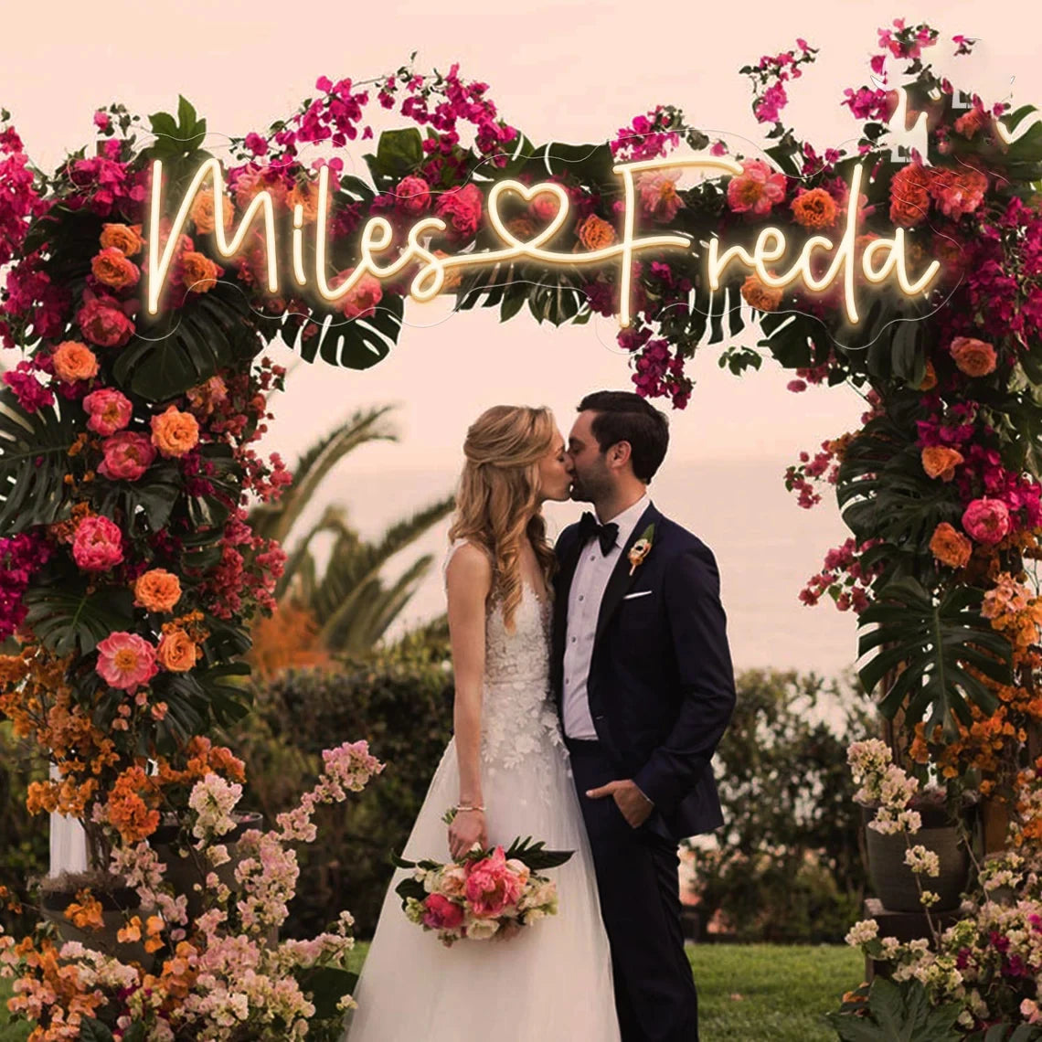 Bride and groom kiss under a floral archway with vibrant pink, orange, and green foliage. Illuminated sign reads "Miles ♥ Freda" against a scenic outdoor backdrop.