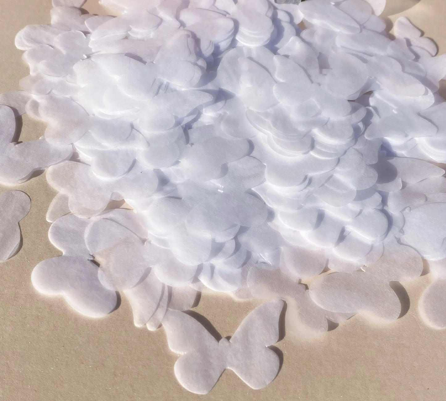 A pile of delicate, white paper butterflies scattered on a light surface, casting soft shadows. The butterflies are intricately cut, showing slight translucency in the sunlight.