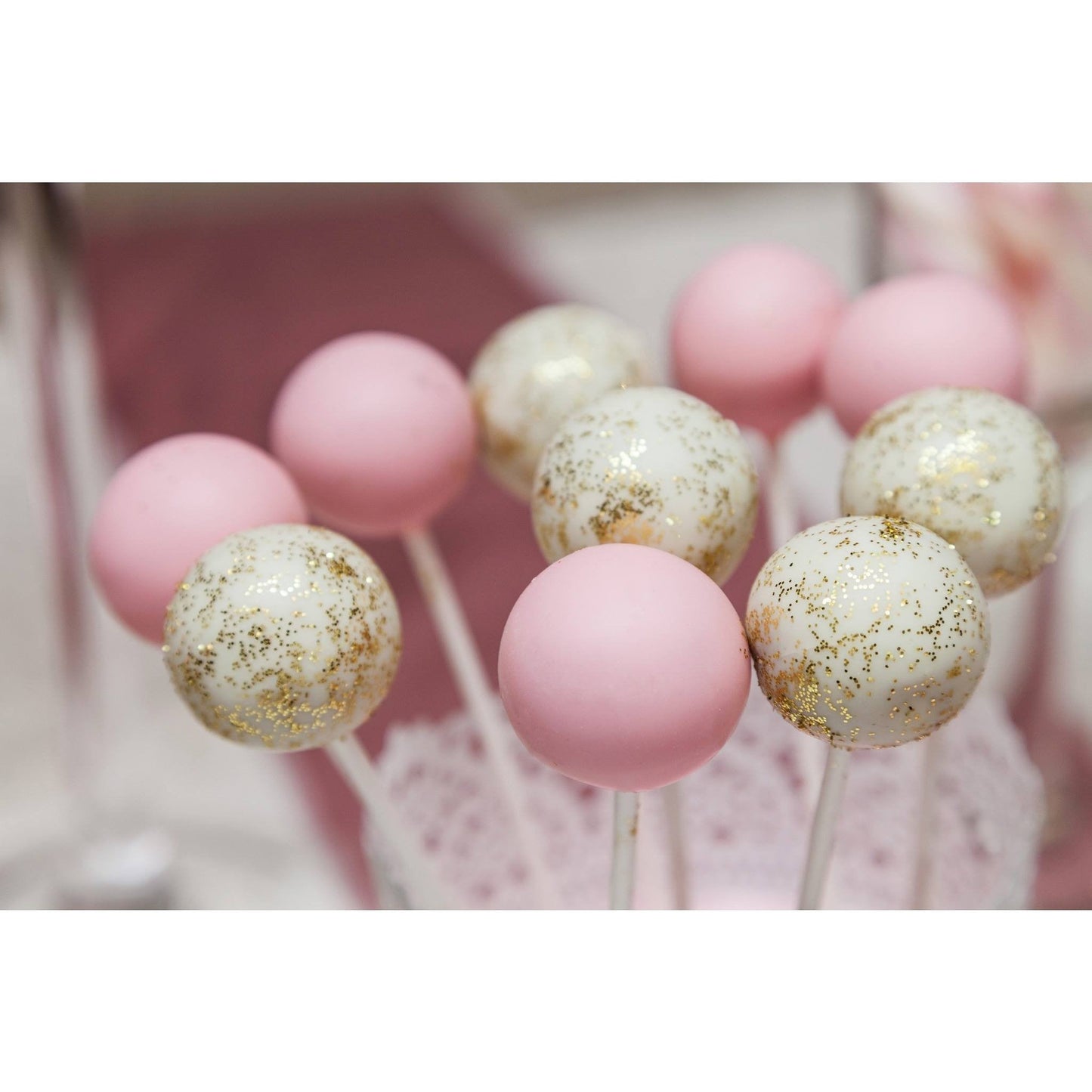 Pink and white cake pops with gold glitter accents arranged in a cup with a decorative doily, set against a soft, blurred background.