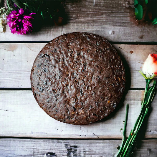 Round chocolate cake on a wooden surface, surrounded by flowers, including a bright pink bloom at the top left and a red and white rose on the right.