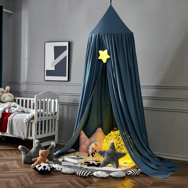 Children's playroom with a hanging dark blue canopy, star decor, cushions, and a glowing ball. A white crib with plush toys is nearby on a wooden floor.