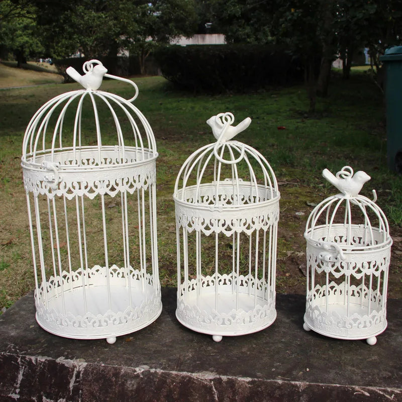 Three decorative white birdcages of varying sizes, each topped with a bird figure, sit on a stone slab in a grassy outdoor setting.