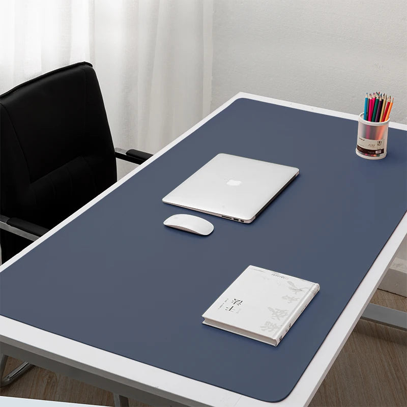 A minimalist desk setup with a navy desk mat, closed laptop, mouse, book, and colorful pencils in a holder. A black chair is placed beside the desk against a light curtain backdrop.