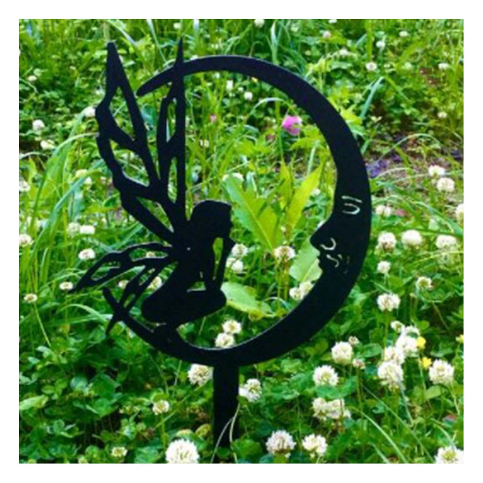 Silhouette of a fairy sitting on a crescent moon garden stake, surrounded by lush green grass and white clover flowers.