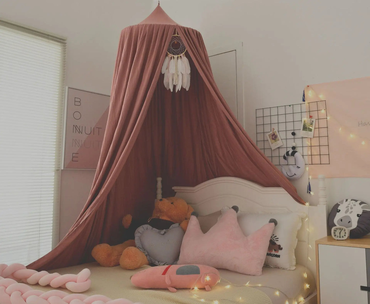 Cozy kids' bedroom with a pink canopy, dreamcatcher, and plush toys on a bed. Walls adorned with fairy lights, photos, and "Bonne Nuit" framed poster. Soft, inviting atmosphere.
