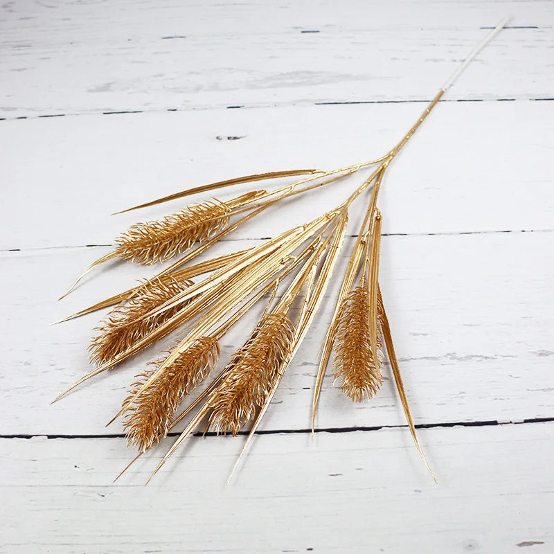 Golden wheat-like stems with textured heads and long, slender leaves are arranged on a white wooden surface.