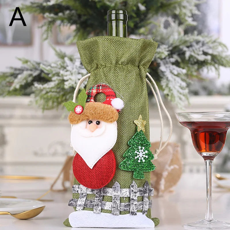 Green fabric wine bag with festive Santa and tree decorations. It's beside a glass of red wine on a table, with a snowy Christmas tree in the background.
