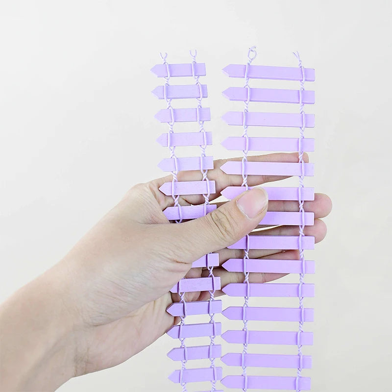 A hand holds two strands of interconnected lavender plastic arrows, resembling a flexible fence or decorative chain, against a plain white background.