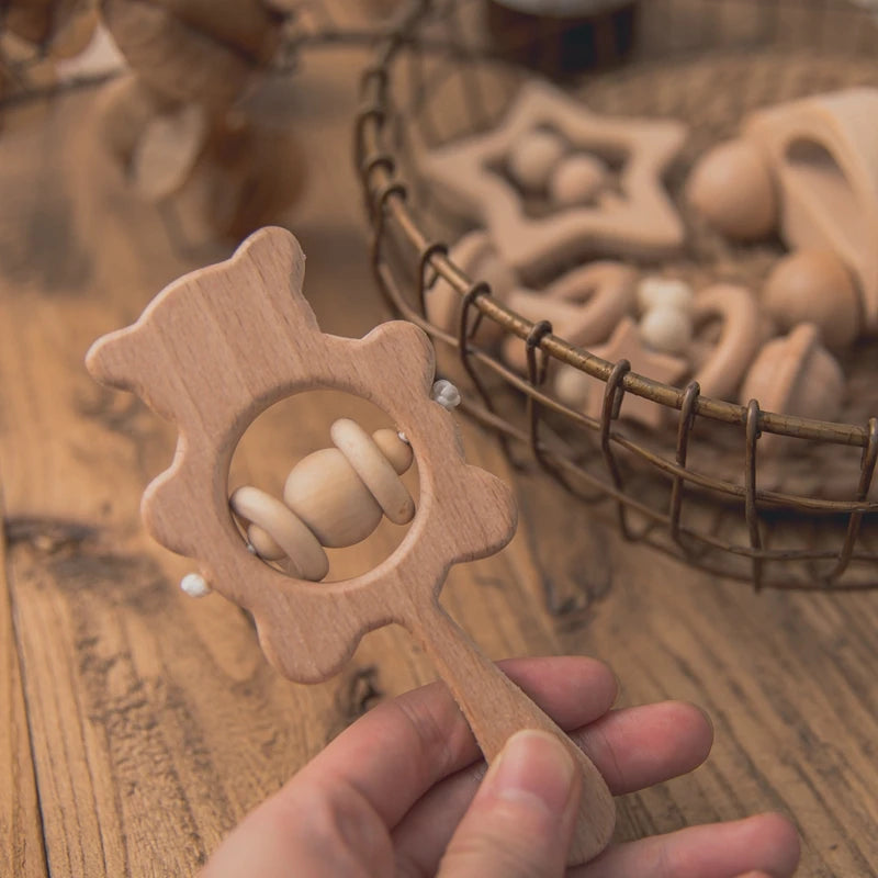 Hand holding a wooden bear-shaped rattle with beads, placed on a wooden table. A wire basket filled with wooden toys is in the background.