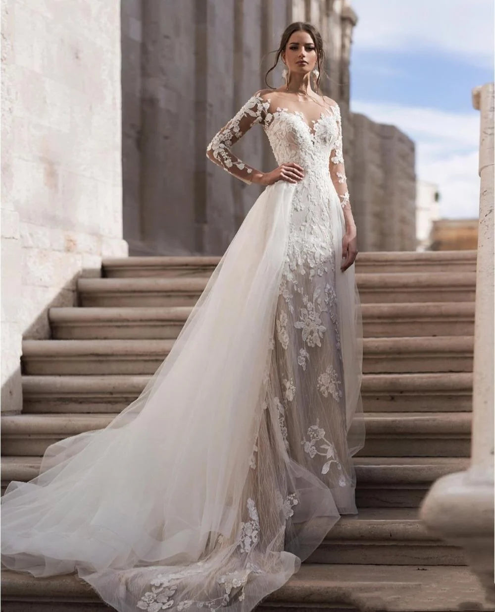 Bride in elegant lace wedding gown with long sleeves and a flowing train, stands confidently on stone steps against a backdrop of light-colored walls and clear sky.