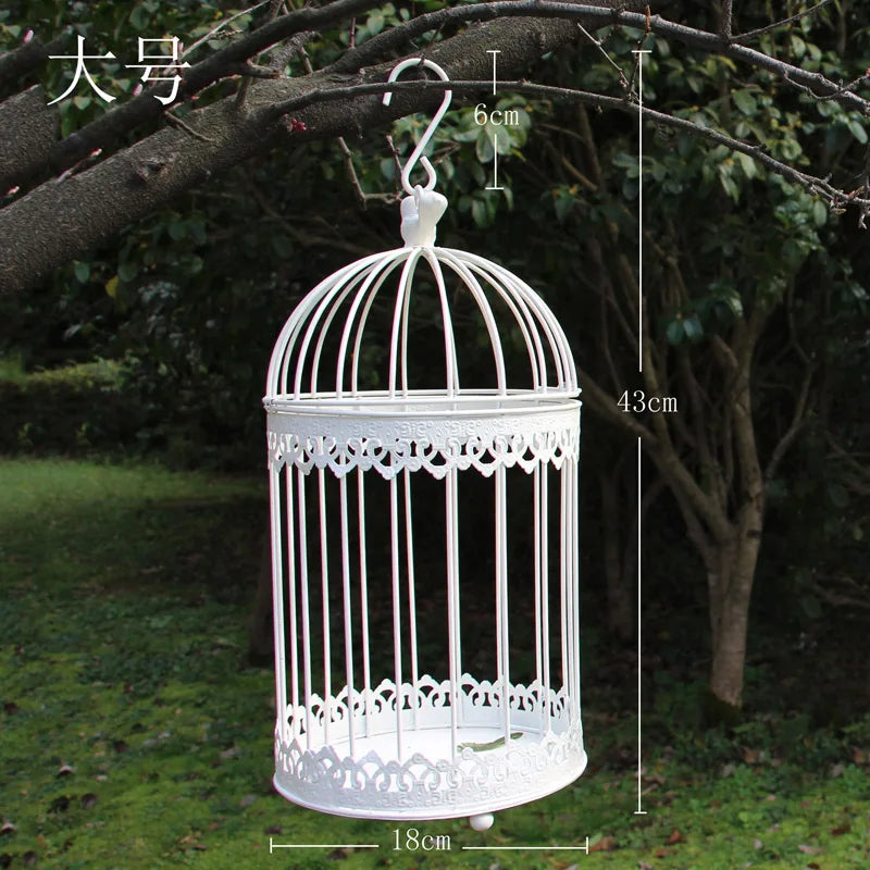 A white, decorative birdcage with a hook is hanging from a tree branch. It measures 43cm in height, 18cm in diameter, and the hook height is 6cm. The cage is surrounded by green foliage.