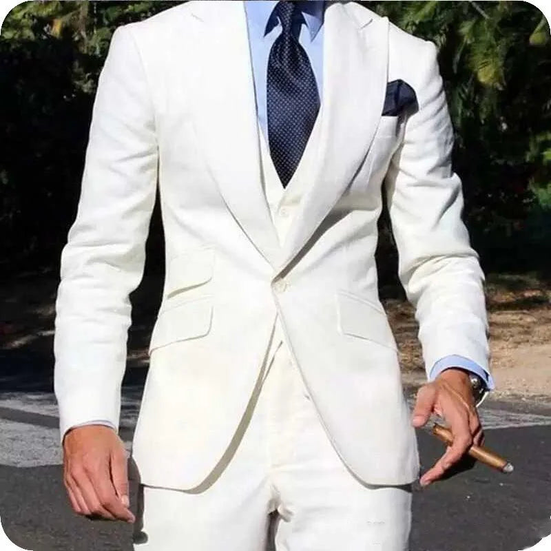 Man in a white suit and blue tie, holding a cigar. The suit features a pocket square, worn over a light blue shirt. Background has greenery and sunlight.
