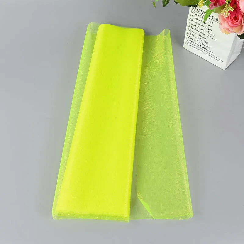 A folded sheet of vibrant neon yellow-green organza fabric lies on a gray surface beside a white paper bag with pink flowers and printed text.