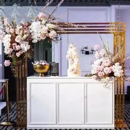 Elegant wedding backdrop featuring a white cabinet with a gold geometric frame adorned with pink and white flowers. A tiered cake and decorative bowl sit on top.