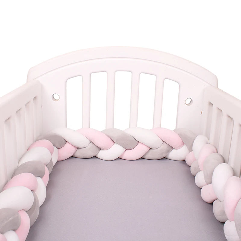 White crib with a soft, braided bumper in pastel pink, gray, and white along the edges, and a gray mattress inside.