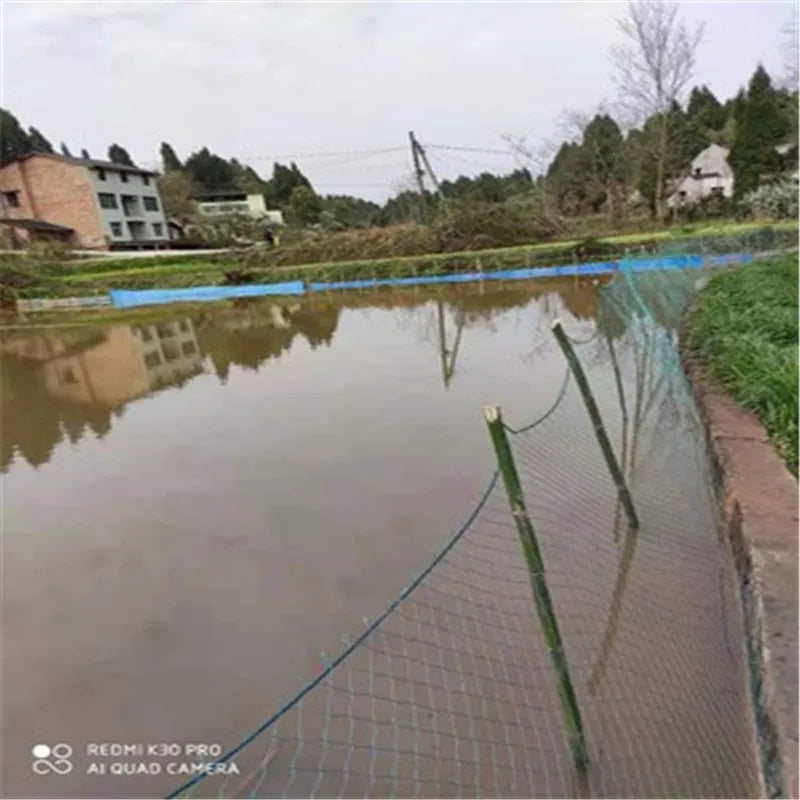 A flooded area with muddy water reflecting nearby houses and trees. A green wire fence leans into the water, with grassy banks visible on the right. The sky is overcast.