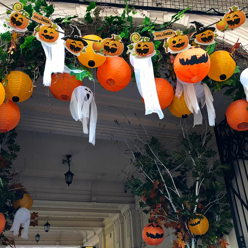 A festive Halloween display features orange lanterns and jack-o'-lantern decorations hanging with small ghost figures in a vine-covered archway. The scene is lively, with contrasting black hats on pumpkins and a backdrop of green foliage and white draped fabric, creating a spooky ambiance.