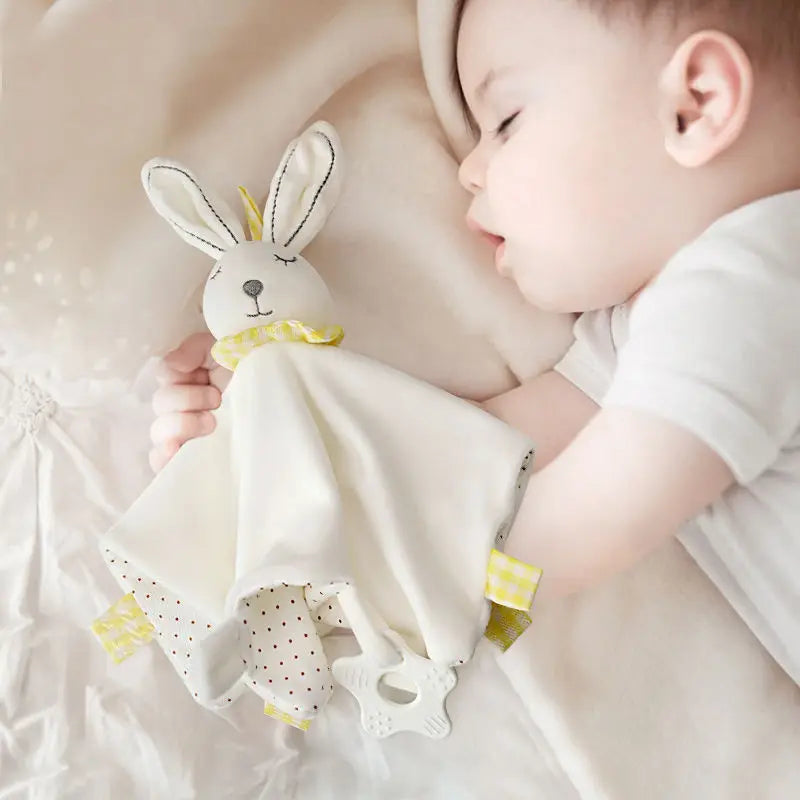 Sleeping baby cuddling a soft plush bunny blanket with yellow and polka-dot patterns on a beige bed. The bunny has floppy ears and the baby wears a white onesie.