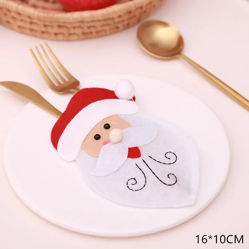 Felt Santa Claus cutlery holder on a white plate, featuring a red hat and white beard, with a golden fork and spoon nearby. Basket and red apple in the background. Size noted: 16*10CM.