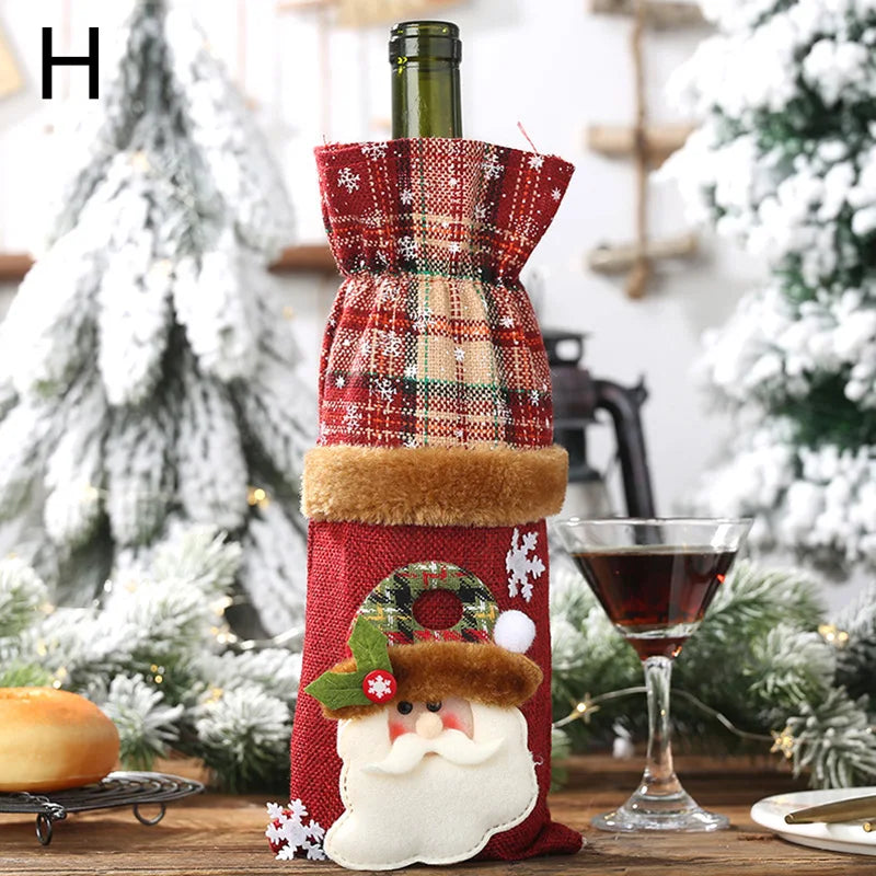 Festive wine bottle with a red plaid Christmas cover featuring a Santa applique. Set on a wooden table with greenery, a pastry, and a glass of red wine, snowy decor in background.