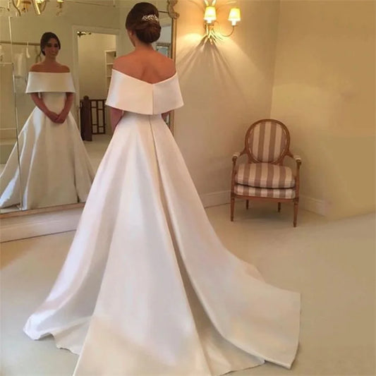 A woman in an elegant off-the-shoulder white gown with a long train stands in front of a mirror. Her hair is styled in an updo with a decorative accessory.
