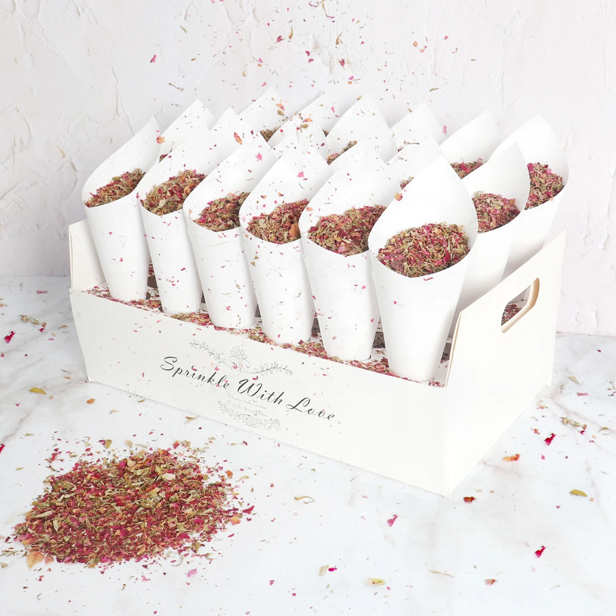 A white box with "Sprinkle With Love" holds 13 cones filled with dried pink and green flower petals. Loose petals are scattered around on a white surface.