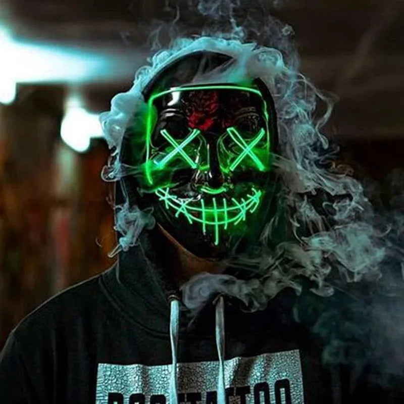 Person wearing a black hoodie and a glowing green LED mask with a stitched smile and X-ed eyes, surrounded by swirling smoke in a dimly lit environment.