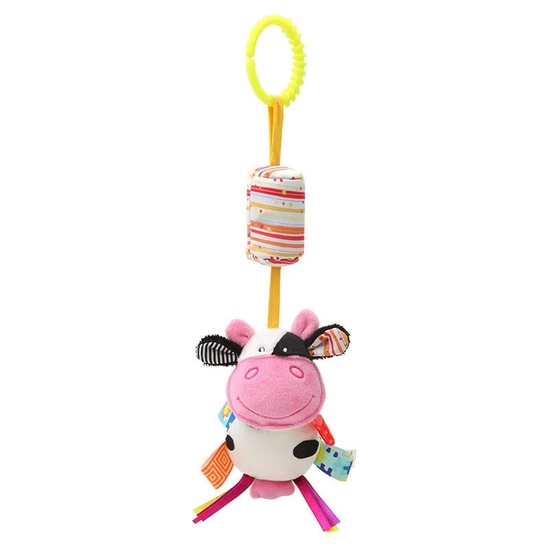 Colorful plush cow baby toy with a smiling face, spotted body, and textured ears. Attached by a yellow loop, it features colorful ribbons and a cylinder with stripes above.