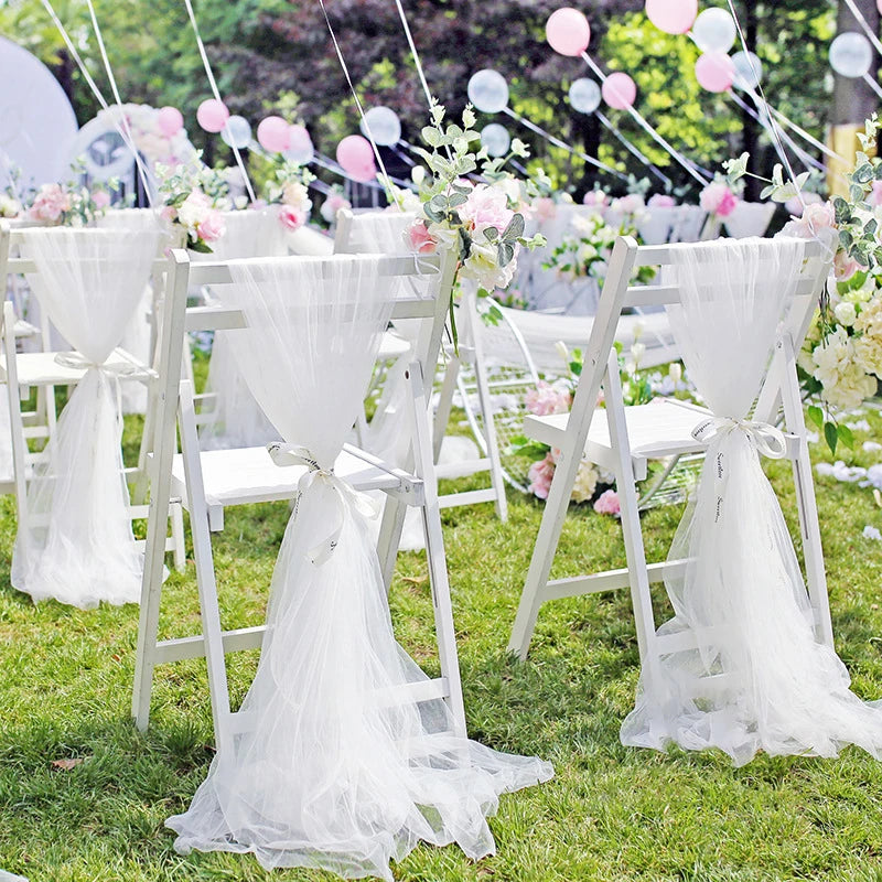 White folding chairs adorned with sheer fabric and pink flowers in an outdoor wedding setup. Pink and white balloons float above on a sunny day.