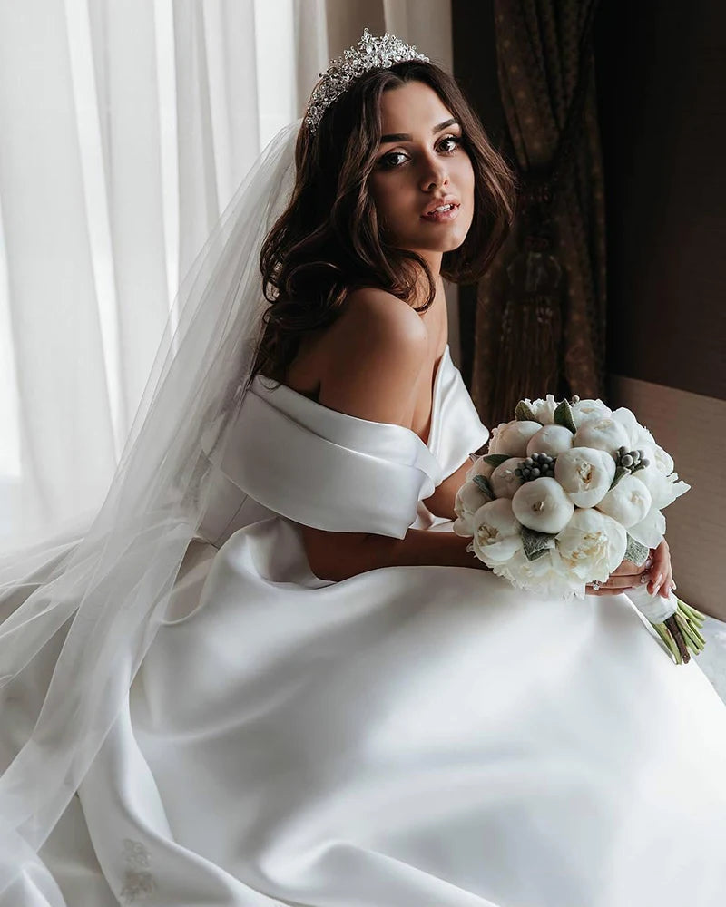 Bride in white satin gown and tiara, holding a bouquet of white flowers, sits by a window with sheer curtains, looking over her shoulder with a serene expression.
