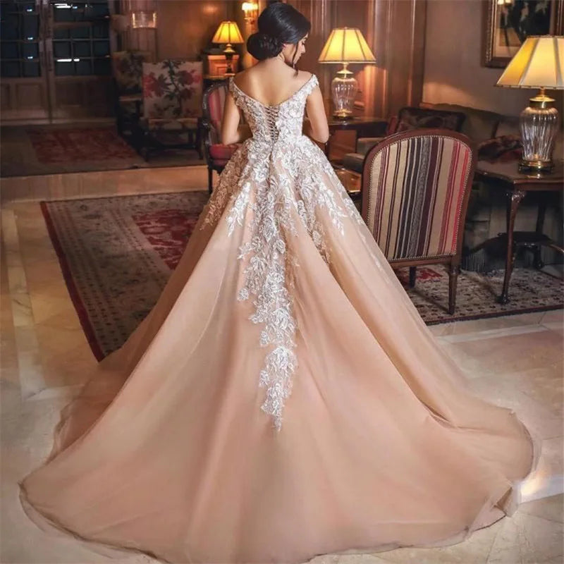 A woman in a luxurious room wears an elegant, off-shoulder ball gown with intricate lace embroidery and a flowing train. The room is softly lit, creating a warm, inviting atmosphere.