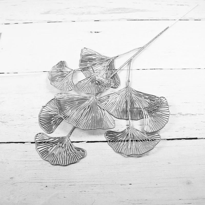 Silver wire art depicting intricate ginkgo leaves arranged on a white wooden surface, showcasing delicate vein patterns in an elegant and minimalist style.