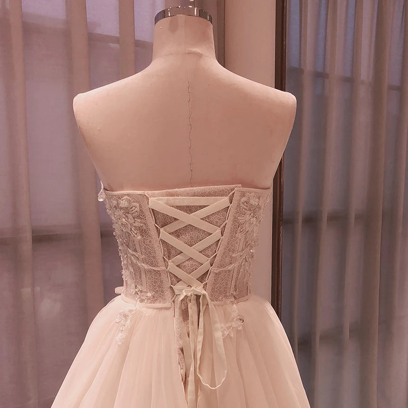Back view of a mannequin wearing a strapless lace-up wedding dress with floral lace details and a flowing tulle skirt, set against a softly lit background with sheer curtains.