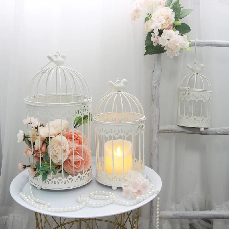 White birdcages with a candle and pink flowers sit on a table with pearl strands. A small birdcage hangs from a wooden ladder, adorned with more pink flowers.