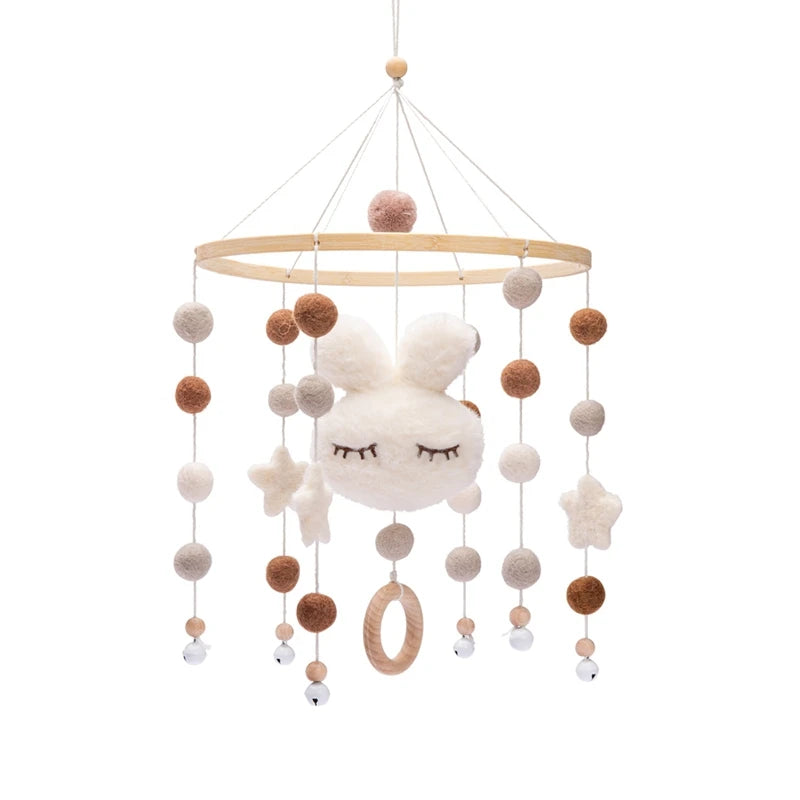 A baby mobile with a wooden ring, felt balls in brown and cream, and a plush bunny with closed eyes. Hanging stars, a wooden ring, and small bells complete the design.