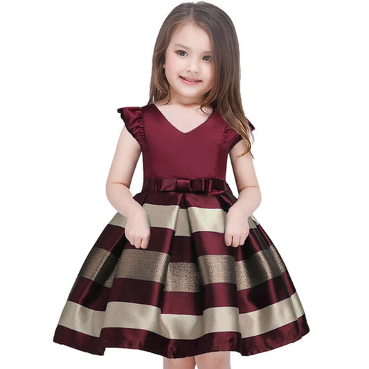 Young girl smiling in a burgundy and beige striped dress with ruffled shoulders and a bow at the waist, standing against a plain white background.