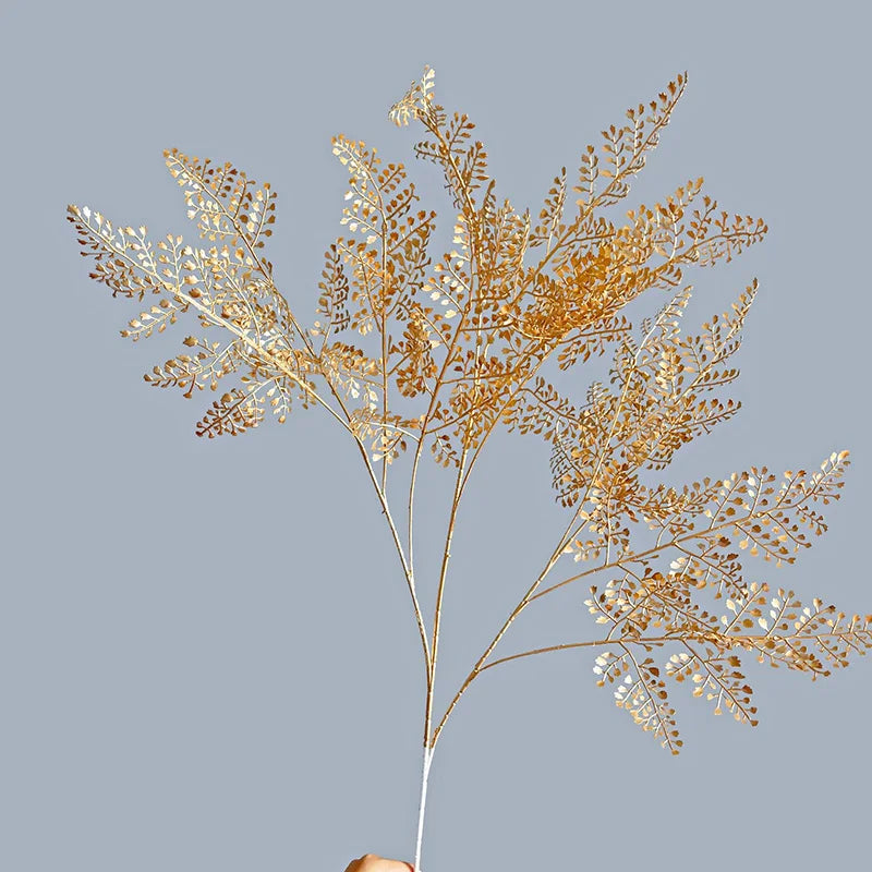A hand holds a delicate, golden botanical branch with intricate leaves against a plain gray background, capturing a minimalist and elegant aesthetic.