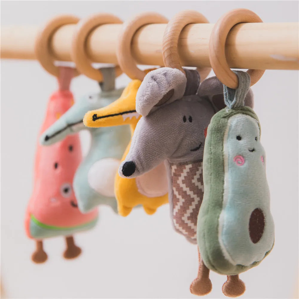 Plush toys shaped like fruit and animals, hanging on a wooden bar by wooden rings. Includes a watermelon, fox, mouse, and avocado, each with cute, embroidered faces.