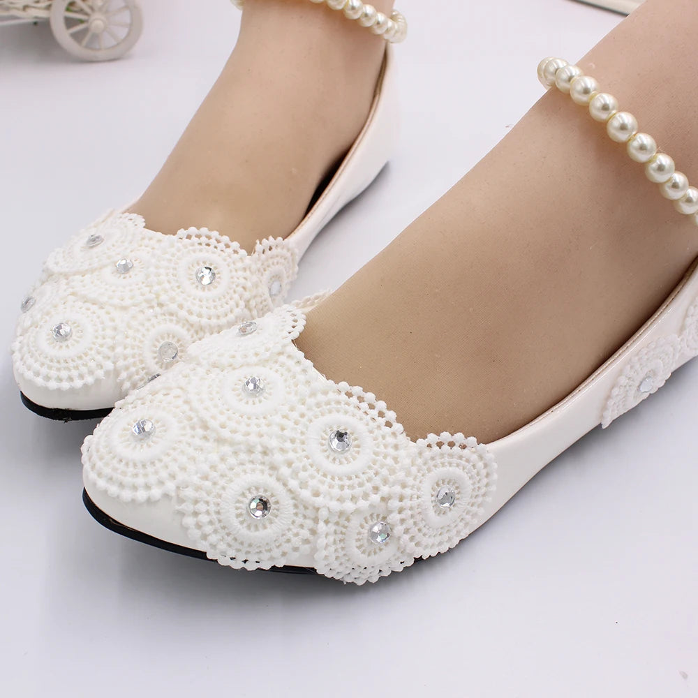 Elegant white flats adorned with intricate lace patterns and small gemstones, paired with a pearl anklet on a light background.