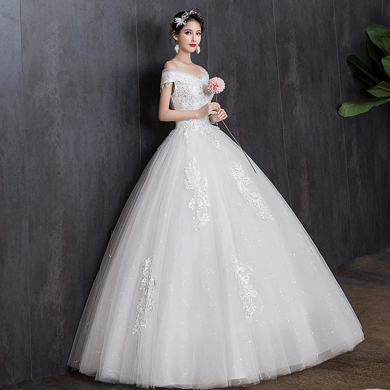 Bride in a white, off-shoulder, lace-embellished gown with a full tulle skirt, holding a pink flower, stands against a dark textured wall. Hair adorned with floral accessories.