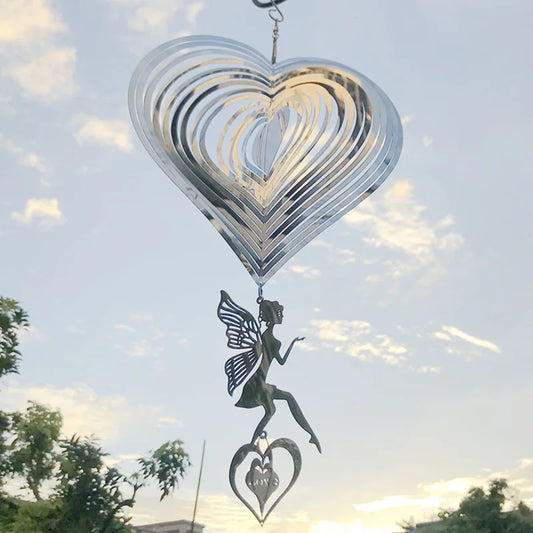 A metallic heart-shaped wind spinner with a dangling fairy silhouette and a smaller heart inscribed with "love" hangs against a cloudy sky with tree silhouettes below.