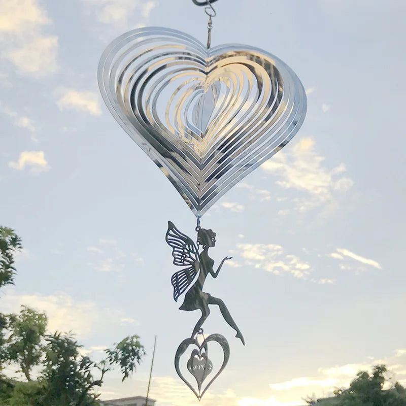 Metallic heart wind spinner with a dangling fairy and heart charm, set against a bright sky with scattered clouds and silhouetted trees in the background.