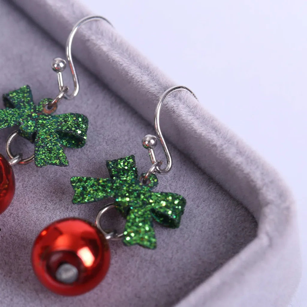 Close-up of festive earrings with glittery green bows and shiny red bells resting in a soft gray jewelry box.