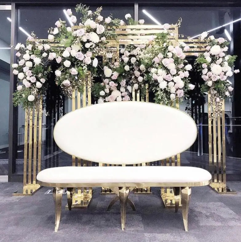 Elegant white and gold bench in front of ornate floral backdrop with white and pink roses, arranged on gold stands.