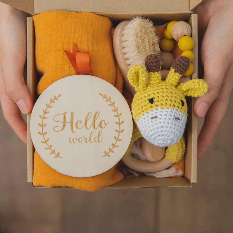 A gift box containing a yellow knitted giraffe toy, a wooden brush, a yellow cloth, and a circular wooden plaque reading "Hello world," all held by two hands.