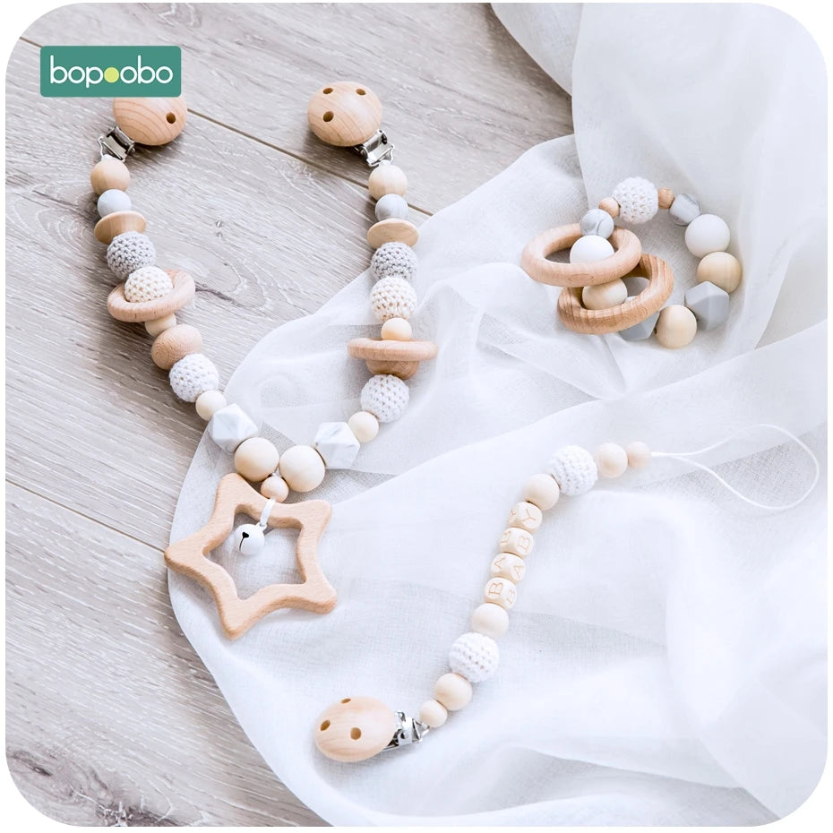 Wooden baby toys on a soft white cloth include a star-shaped teether, a pacifier clip with "BABY" beads, and a ring rattle, all with natural and white colors.