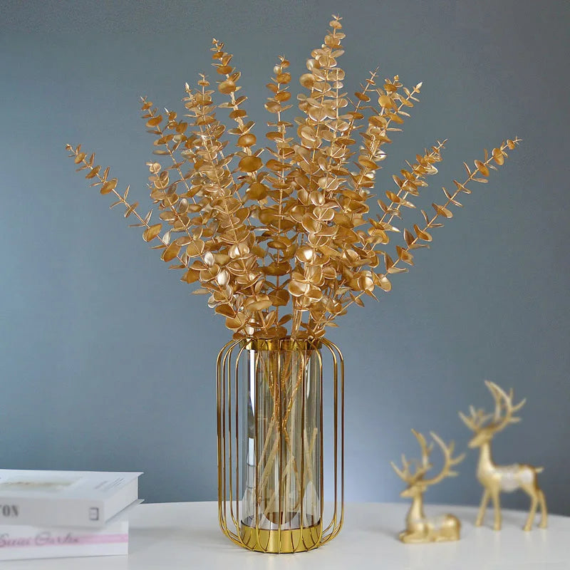 Golden eucalyptus branches in a metal-framed glass vase on a white table with two gold deer figurines and books against a gray background.