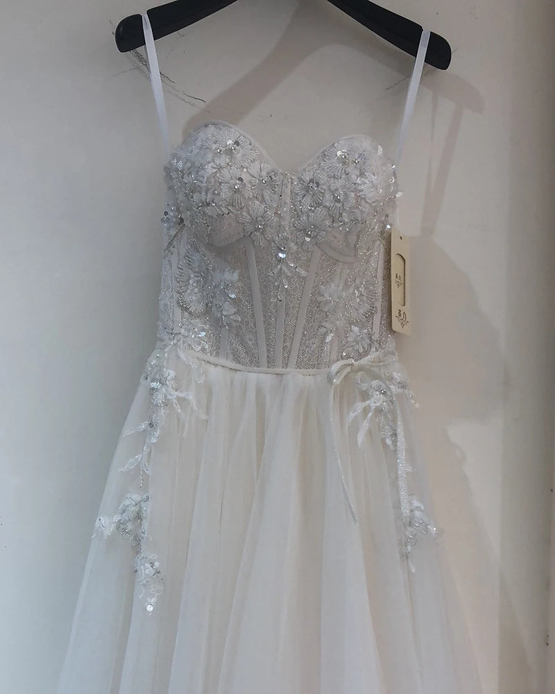 Elegant strapless white gown with a fitted, intricately beaded bodice and a flowing tulle skirt. Delicate floral appliques accentuate the waist and skirt, displayed on a hanger.