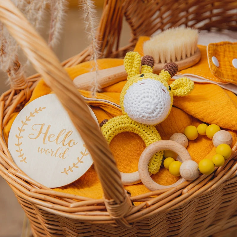 Wicker basket with a yellow giraffe toy, baby brush, yellow blanket, wooden bead toy, and a round sign that reads "Hello world" on an orange cloth background.