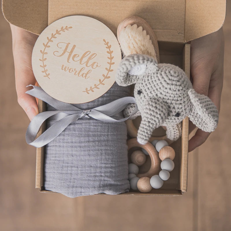 Gift box with a "Hello world" sign, gray fabric tied with a ribbon, a crocheted elephant toy, a baby brush, and a wooden bead bracelet. Hands hold the open box.